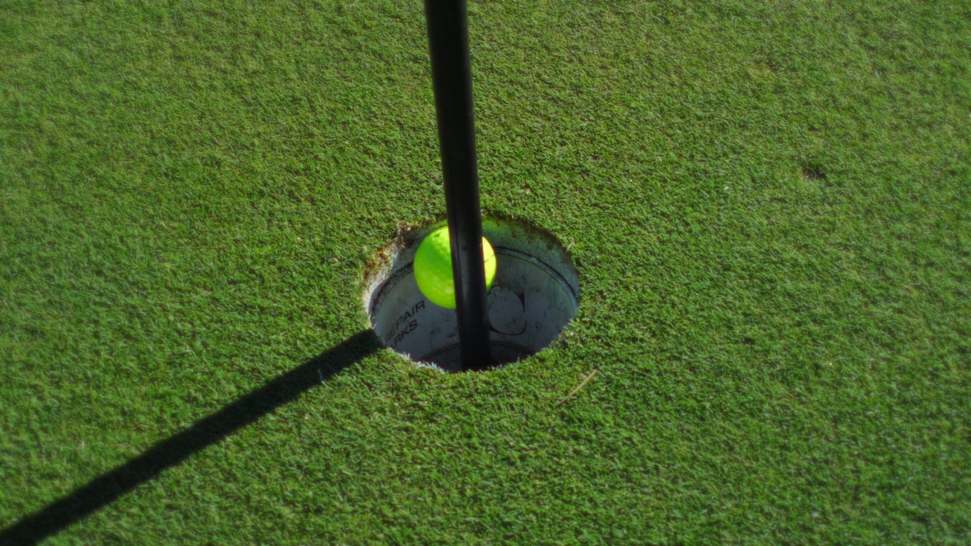 Neongul golfboll i hål bredvid flaggan på en solig green.